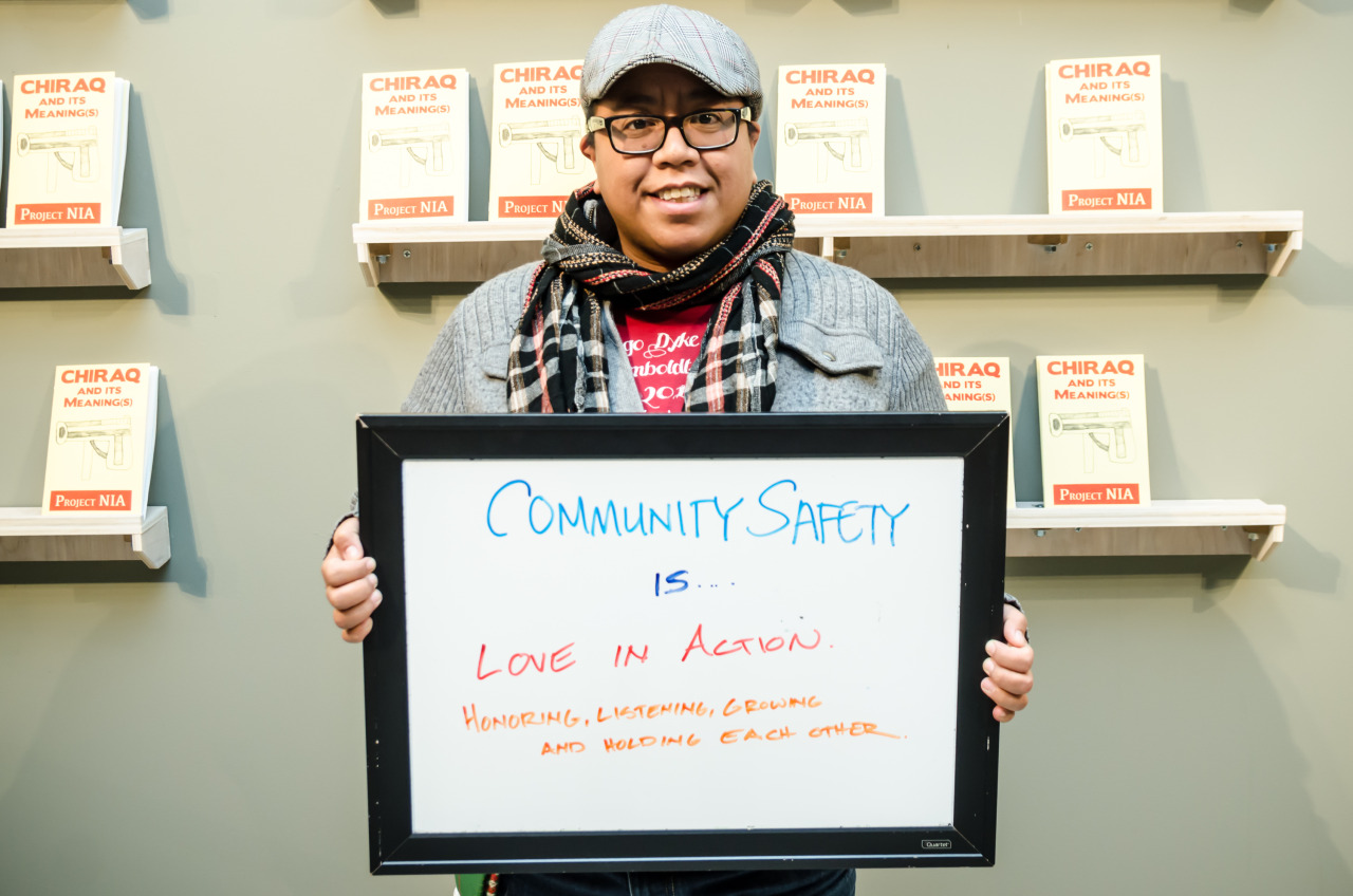 Image: A person of color stands in front of a wall with shelves that have pamphlets with the words “CHIRAQ And Its Meaning(s)” by Project NIA. This person holds a dry-erase board with “Community Safety Is… Love in Action. Honoring, Listening, Growing, and Holding Each Other.” in the center. Photo courtesy of the artist.
