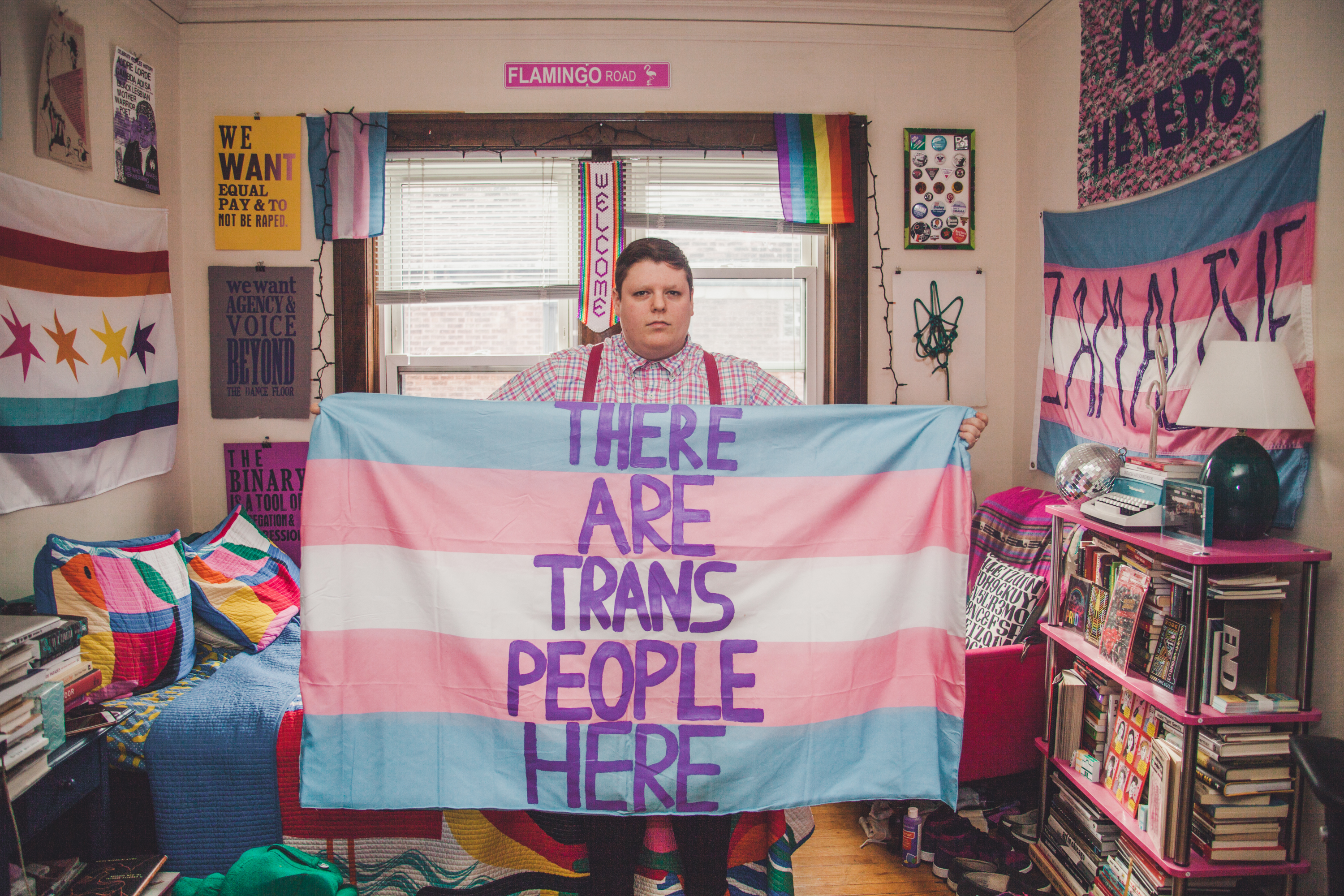 Image: H. Melt holds open a blue, pink, and white banner, the transgender pride flag, that says "There are trans people here" in purple letters in their apartment. Their bed, a bookshelf, and several other printed banners and signs can be seen in the background. Photo by Ally Almore.