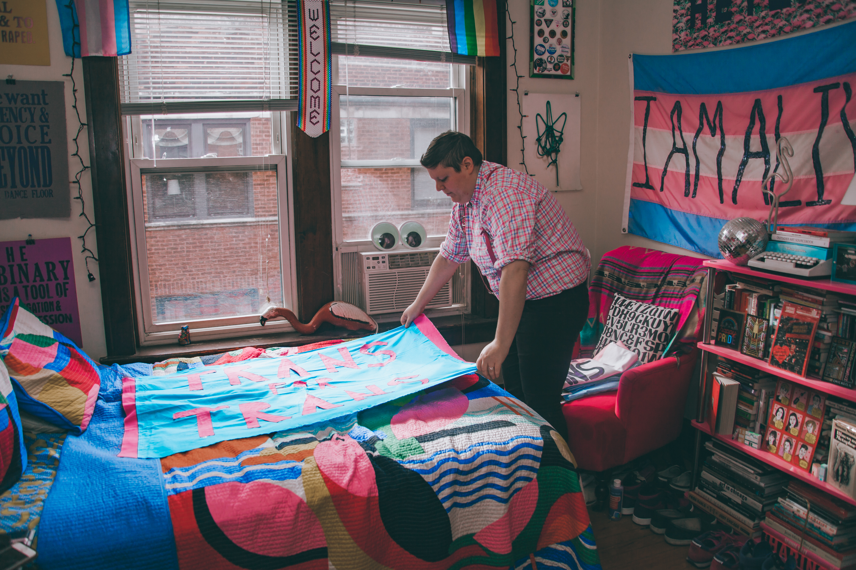 Image: H. Melt arranges a blue banner that says "Trans for Trans" on a bed with a multicolored bedspread in their apartment. Photo by Ally Almore.