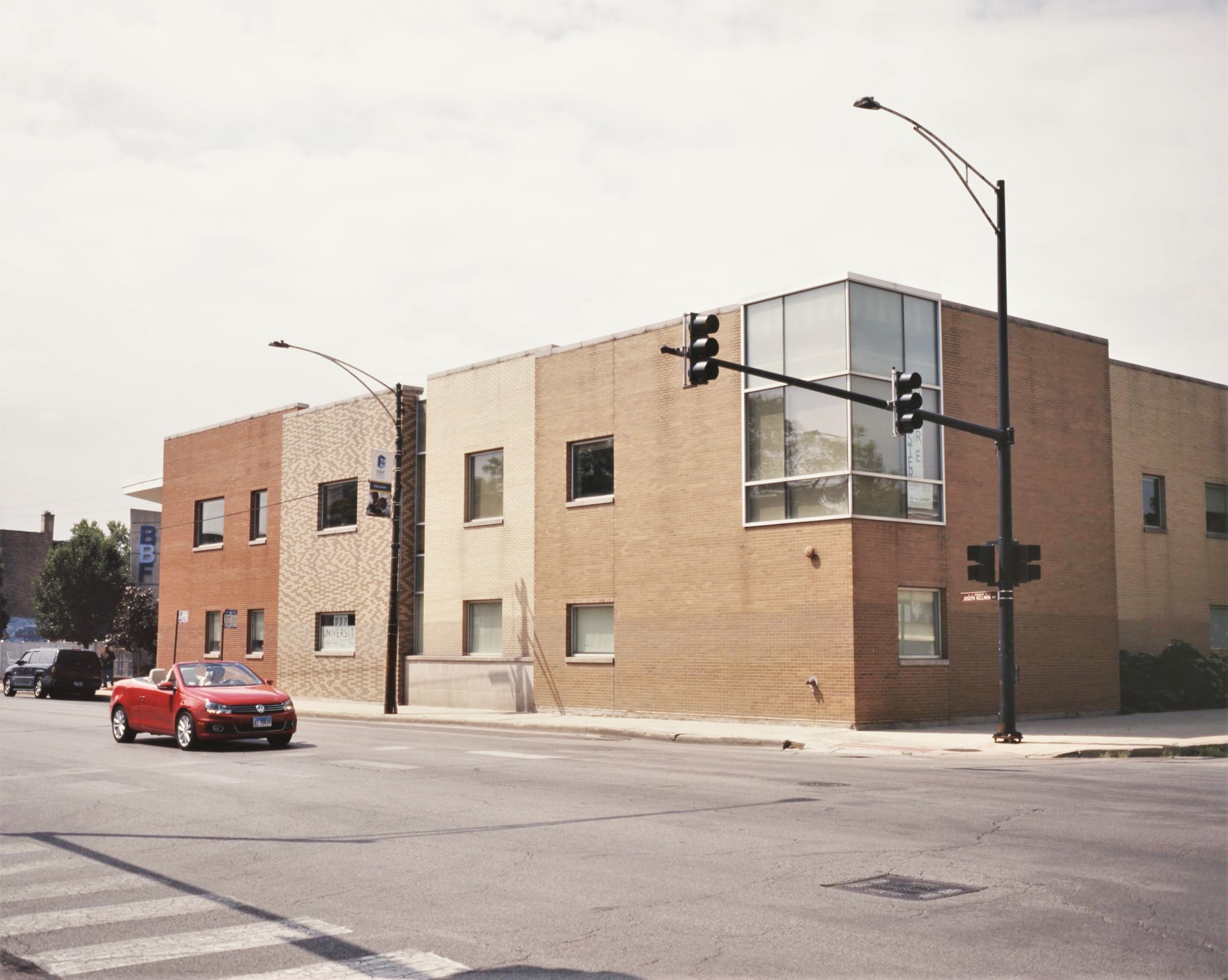 Image: The BBF Family Services Center, seen on a sunny day from the NE corner of 15th and Pulaski in North Lawndale. Designed by architect Lucien Lagrange, the building’s long, two-story play-block facade varies in dimension and brick pattern from end to end. A red Volkswagen cabriolet with the top down is stopped momentarily at the light in front of the building. Photo by Eric K. Roberts.