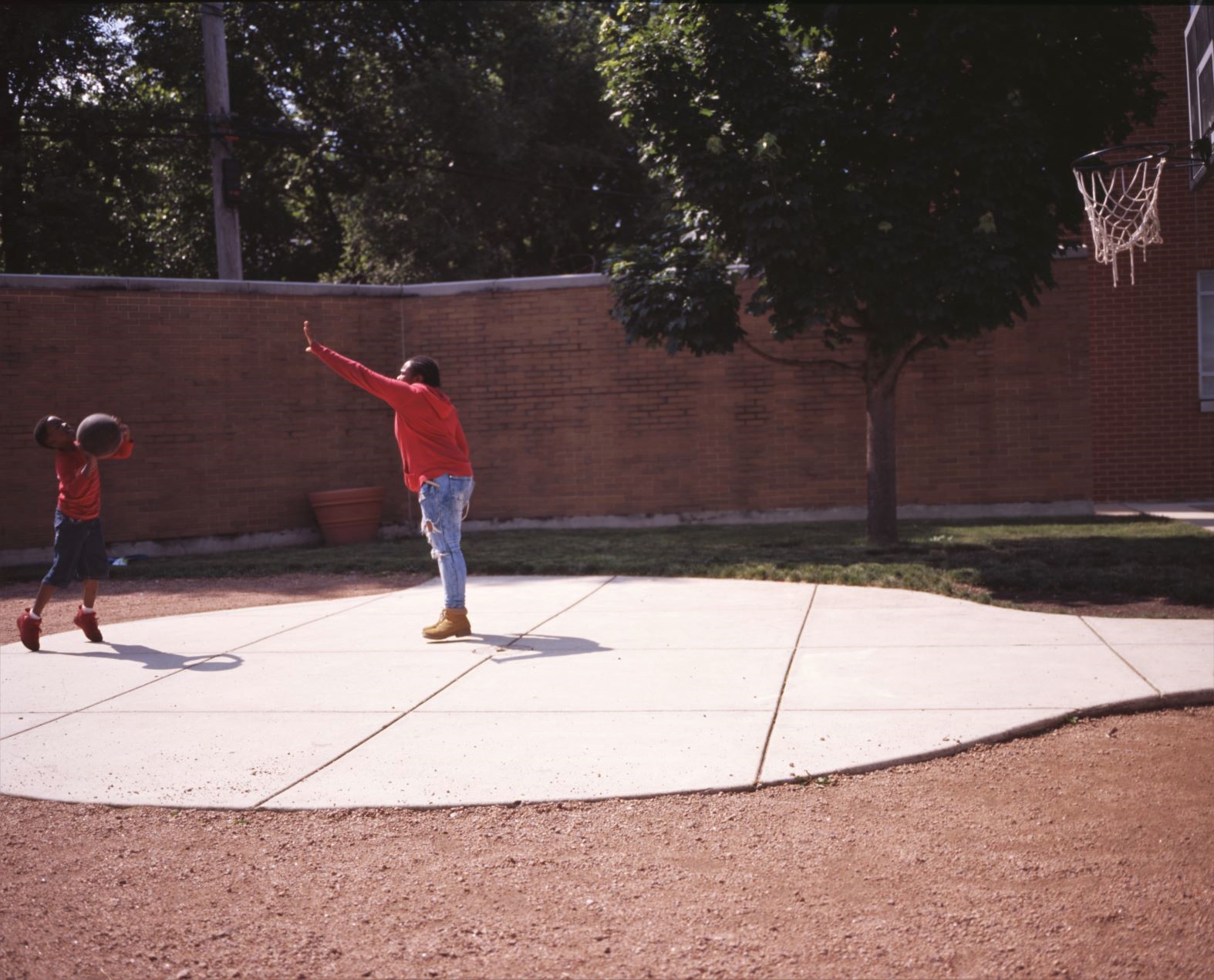 Image: A child in a long-sleeved red t-shirt and matching gym shoes shoots a jumper over the outstretched arm of a slightly older child in a red hoodie and wheat boots in the courtyard of BBF Family Services. Photo by Eric K. Roberts.