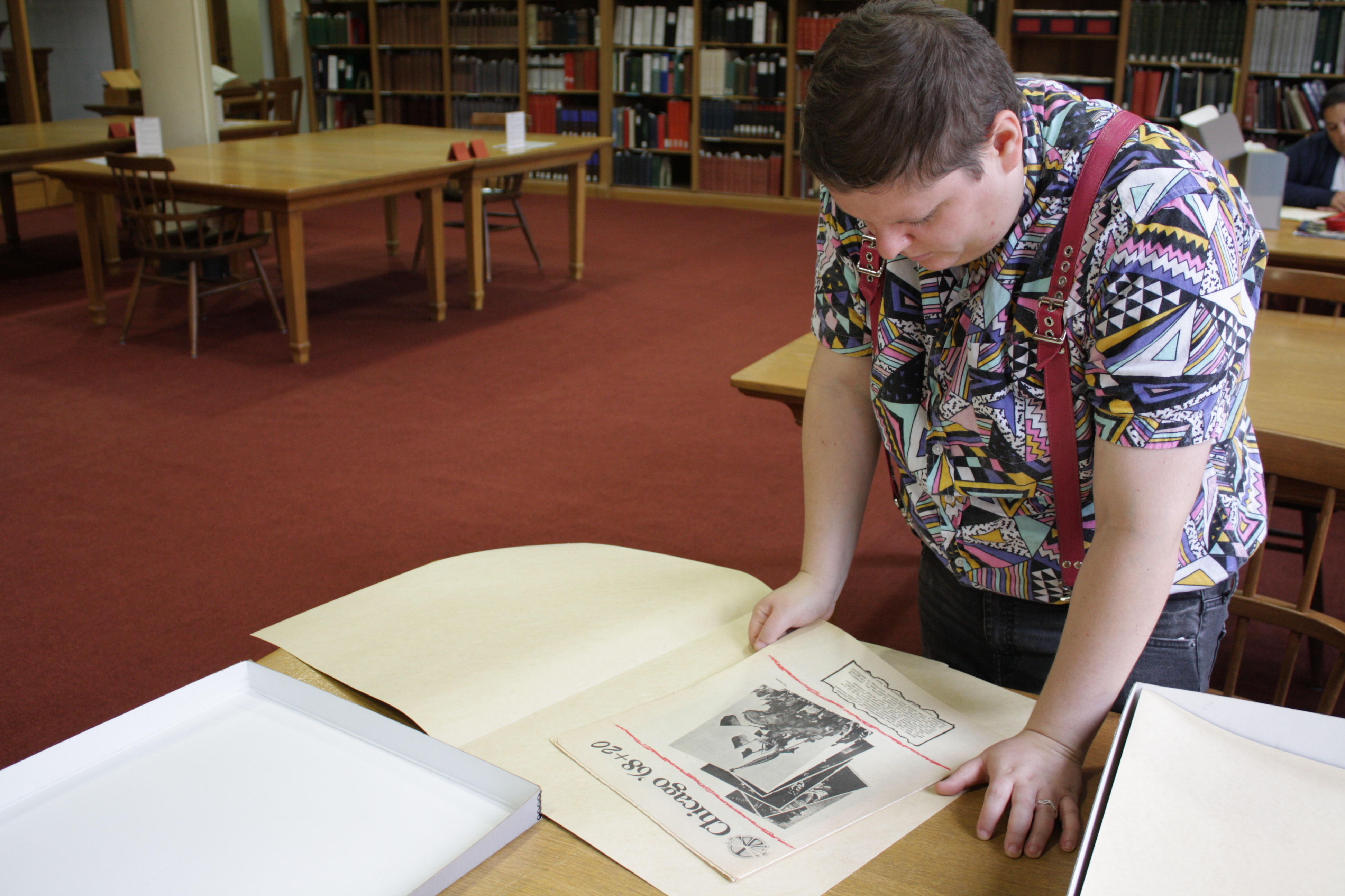 Image: H. Melt stands over a table at the Newberry Library looking at a piece of ephemera in an archival folder. They are wearing a shirt with brightly colored geometric designs and red suspenders. Photo by Ally Almore.