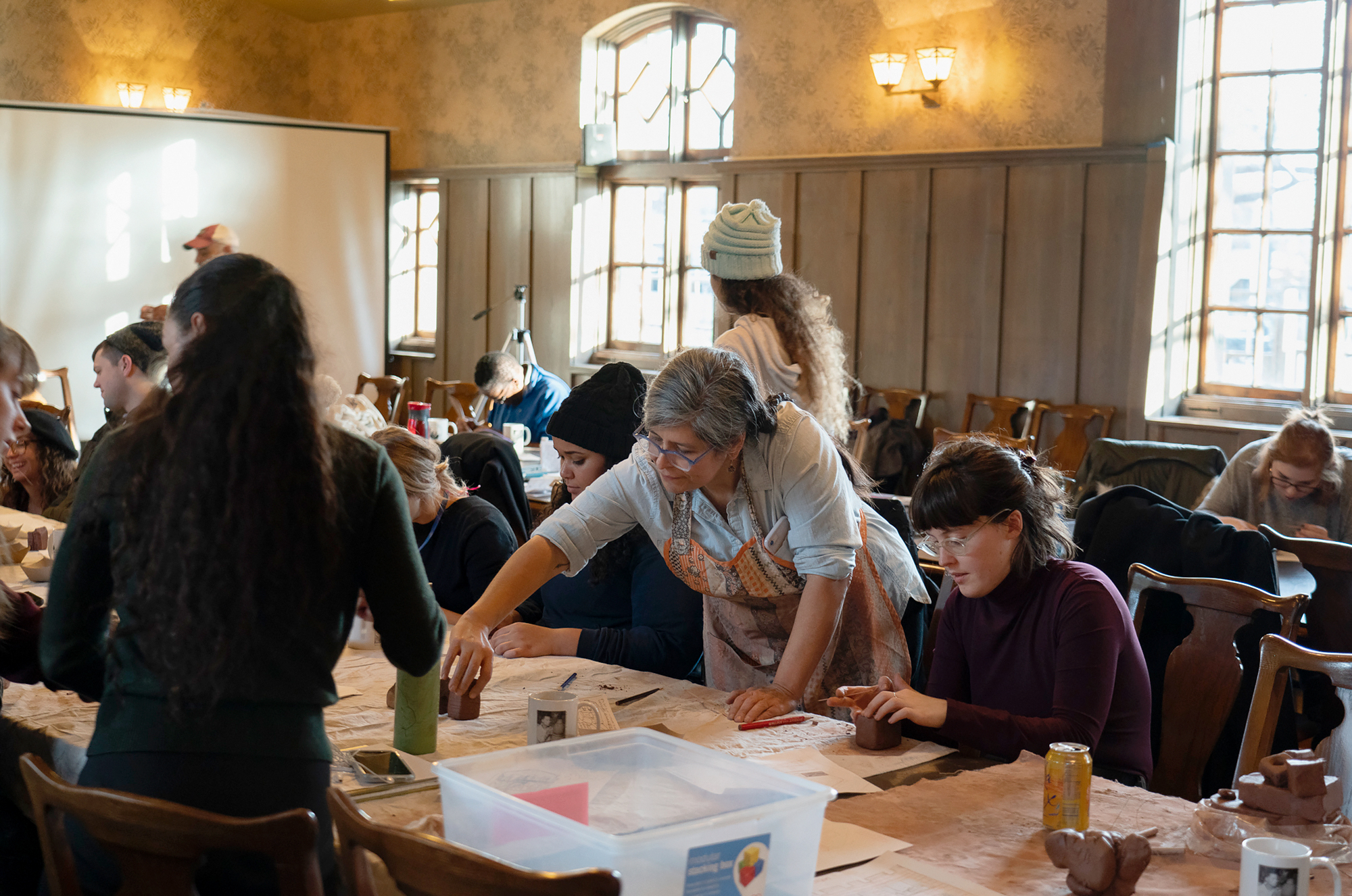 Image: Nicole Marroquin (standing at center, with shirt sleeves rolled up) leads a ceramics workshop at Hull House in November. She is surrounded by participants sitting at tables molding clay into the shapes of buildings. Photo courtesy of Jane Addams Hull-House Museum / Jesse Meredith.