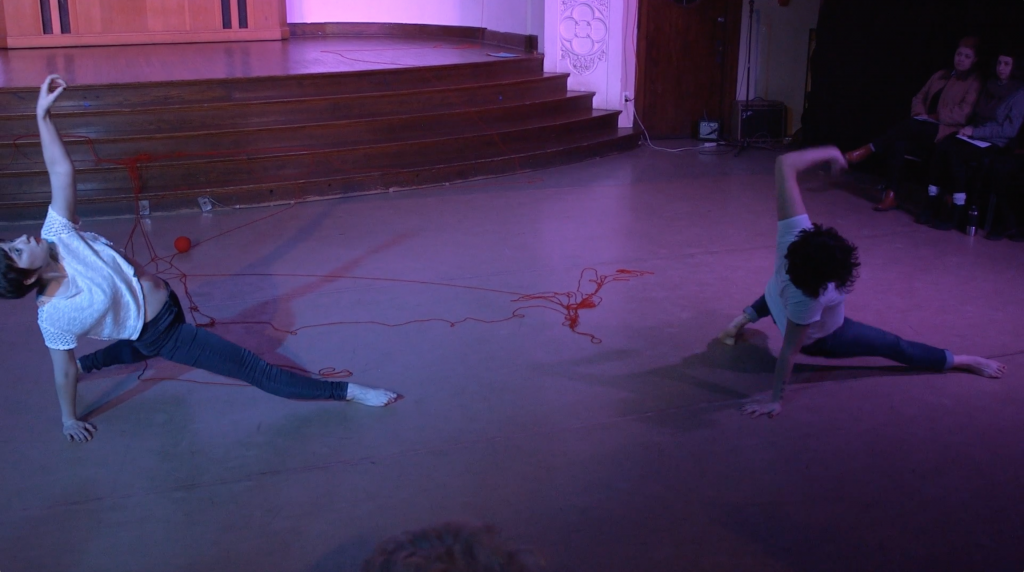 Image: Maggie Robinson and Allison Sokolowski performing in “I Am.” The dancers suspend themselves off the floor in variations of the same pose, as if pausing while sliding into a split. Allison reaches and looks up at the ceiling and Maggie reaches and looks toward the audience. Around them on the floor are strands of red yard. The lighting is dim and cool, with purple highlights. Audience members are slightly visible in the foreground and background. Still from a video by John Borowski.