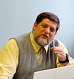 Image: Photo of Arnie Aprill, arts and learning consultant with Envisioning Justice. Arnie Aprill turns toward the camera as he speaks while gesturing with his left hand. He is wearing a light yellow shirt with a gray sweater vest and light colored tie and has a laptop open in front of him. Photo courtesy of Arnold Aprill. 