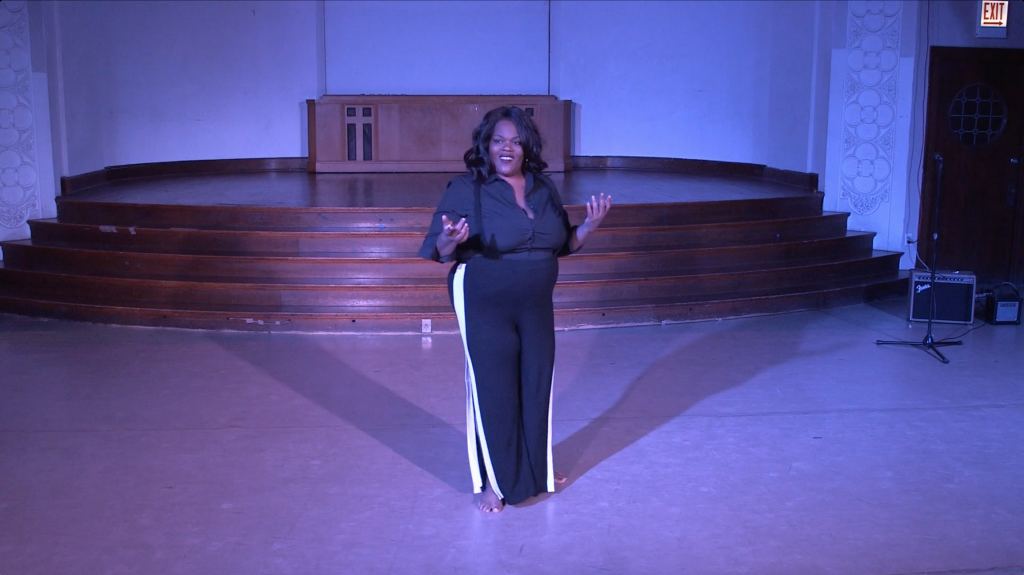 Image: Jae Green performing in “Wax.” Jae smiles brightly at the audience, her arms bent at the elbows with palms facing up. She stands in the middle of the floor, her shadow cast toward the stage in two directions. The lighting is dim and cool (blue-purple). Still from a video by John Borowski.