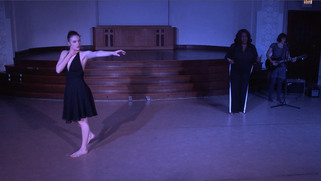 Image: Carly Broutman, Jae Green, and Michelle Shafer performing in “Wax.” In the foreground, Carly steps forward, with one hand on her throat and her other arm stretched across the frame, her face looking that way too. In the back corner, Jae speaks, palms up in a gesture, as Michelle plays electric guitar. The lighting is dim and cool (blue-purple). Still from a video by John Borowski.