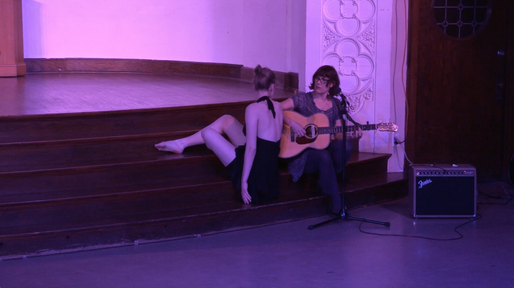 Image: Carly Broutman and Michelle Shafer performing in “Wax.” This slightly closer shot shows Carly and Michelle sitting on the stage steps, turning to look at each other. Carly’s back is to the audience and, though her back is positioned straight-up, her legs are folded or extended onto higher stairs than where she is sitting. Michelle holds an acoustic guitar and sits more commonly, with one foot on a lower step and another on the floor. The lighting is dim and pink-purple. Still from a video by John Borowski.