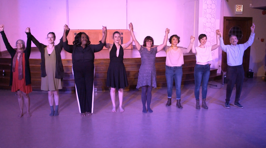 Image: The curtain call, showing all performers from the Body Passages culminating event. The performers stand in front of the stage, smiling and holding hands, arms up in the air. The lighting is pink-purple. Still from a video by John Borowski.
