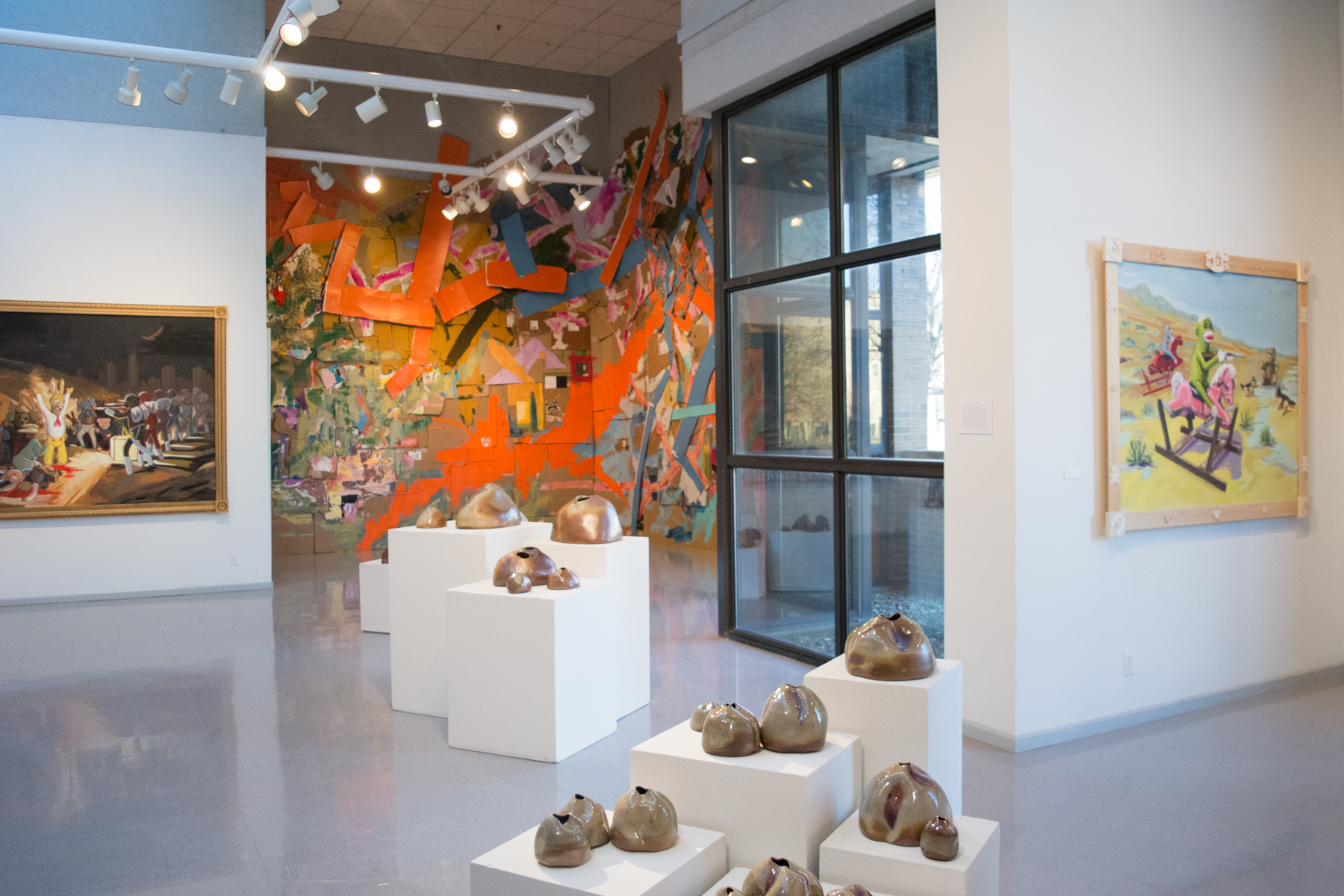 Installation shot of the MA Exhibition in Heuser Art Center at Bradley University. Ceramic pieces by Natalie Zelman are in the foreground. Framed oil paintings on the walls to the left and right by Jack Crouch. A large mixed-media painting installation hangs on the wall in the background. 