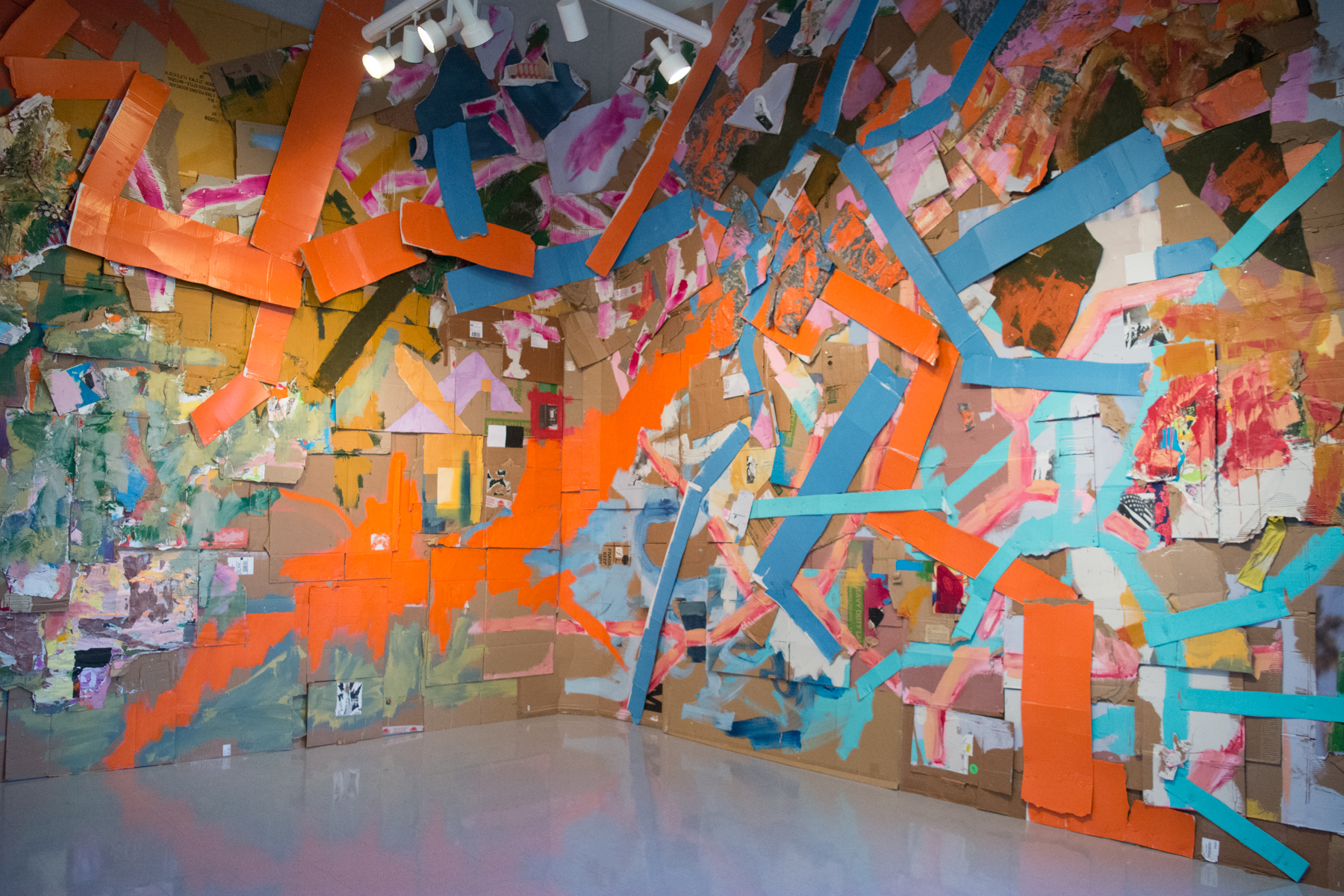 A large floor to ceiling painting installation by Peter Ahart, titled Portrait of an American Solider, hangs in the back of the gallery. The piece is a painting on cardboard, fabric, and collage paper. Orange and blue marks wind through the painting that spans three walls. 