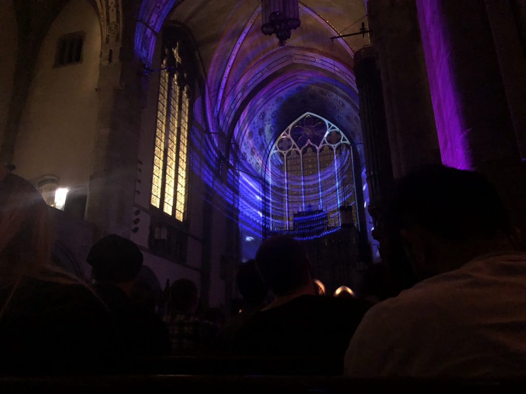 Photo by S. Nicole Lane. Silhouettes of people from the crowd are sitting at the pews. There is a purple circular light shining on the stained glass. To the left, the windows and stained glass have a yellow light. There is a green stream of light on the right side of the image. 