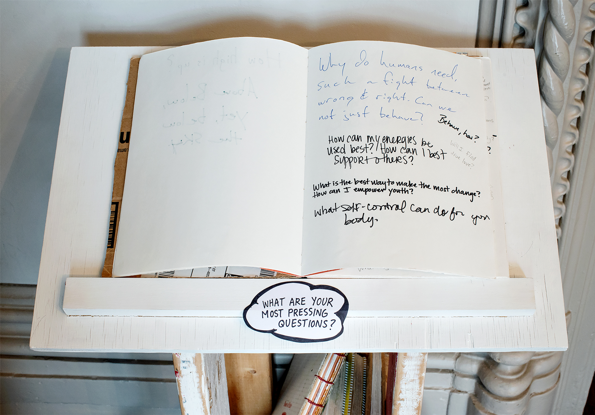 Image: Detail view of a book and prompt set on one of the mobile pedestals in the Everything on Wheels series, placed in one of the downstairs rooms at Hull-House. The prompt says “what are your most pressing questions?”, and people have written answers such as “How can my energies be used best? How can I best support others?”, or “What is the best way to make the most change? How can I empower youth?” Photo by Greg Ruffing.