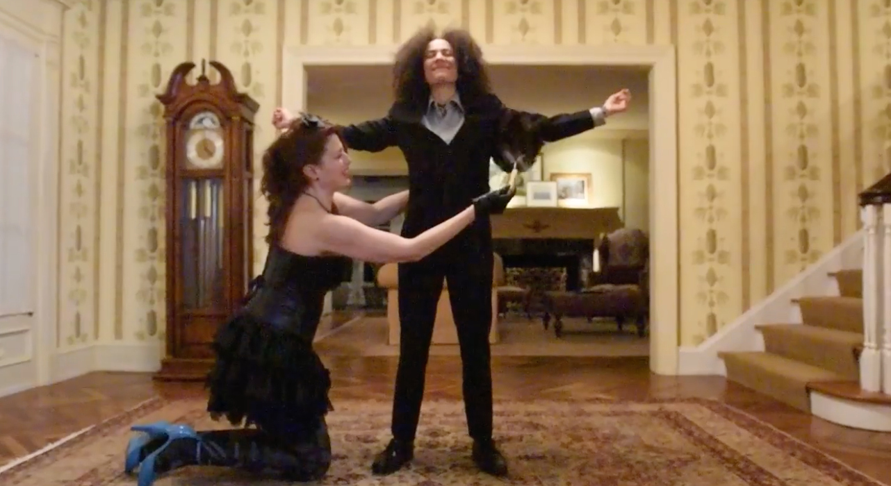 Image: Film still from "Thirst Trap" featuring actors Weezy Carpino (on knees) and Monique Ligons (standing). Here we see Weezy feather dusting Monique in her opulent estate as she prepares for a special dinner date. Photo courtesy of Heather Raquel Phillips.