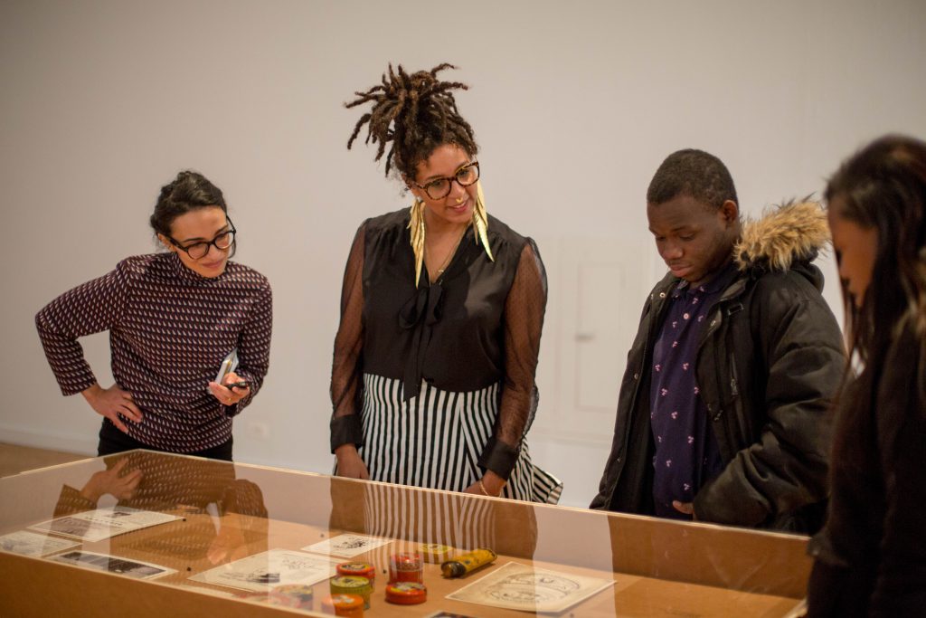 Image: The author, D. Denenge Duyst-Akpem, and two young visitors look at beauty product containers and printed materials displayed in a vitrine in the "African American Designers in Chicago" exhibition at the Chicago Cultural Center. Photo by Ireashia Bennett.