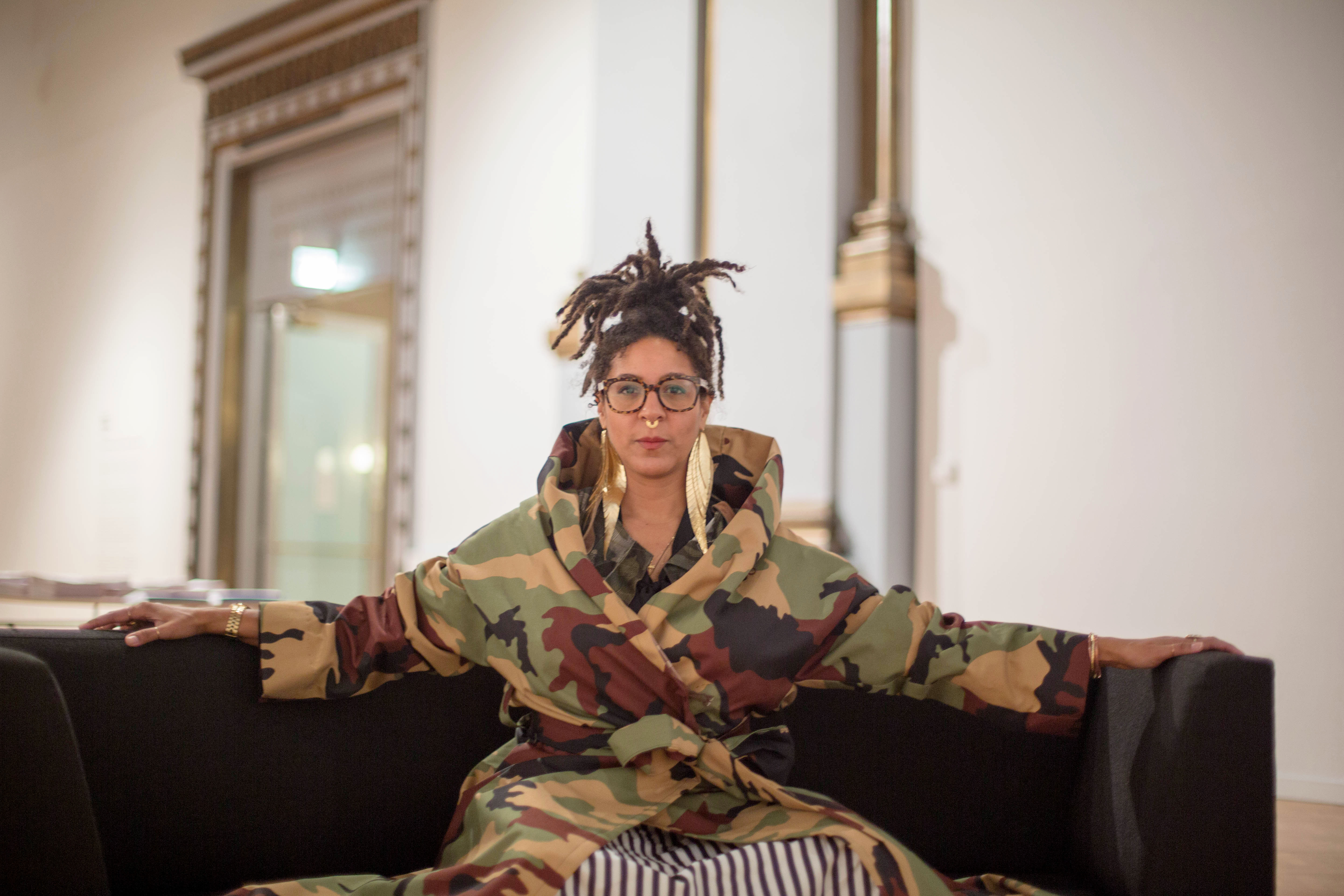 Image: D. Denenge Duyst-Akpem wearing the garment "The Camo Coat, part of the Osanyin Commemorative Project," and sitting on a black sofa in the "African American Designers in Chicago" exhibition at the Chicago Cultural Center. Photo by Ireashia Bennett.