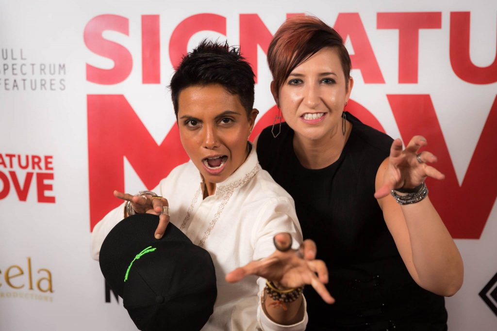 In this medium shot, Fawzia and Lisa pose in front of a white wall emblazoned with the red “Signature Move,” grey Full Spectrum Features, and other logos. Both Fawzia and Lisa look at the camera with playful snarls and with fingers bent like cat claws. Fawzia wears a long-sleeved white button-up and, in one hand, holds a black baseball cap with neon green detailing. Lisa wears a black sleeveless shirt. Both wear multiple bracelets and rings.