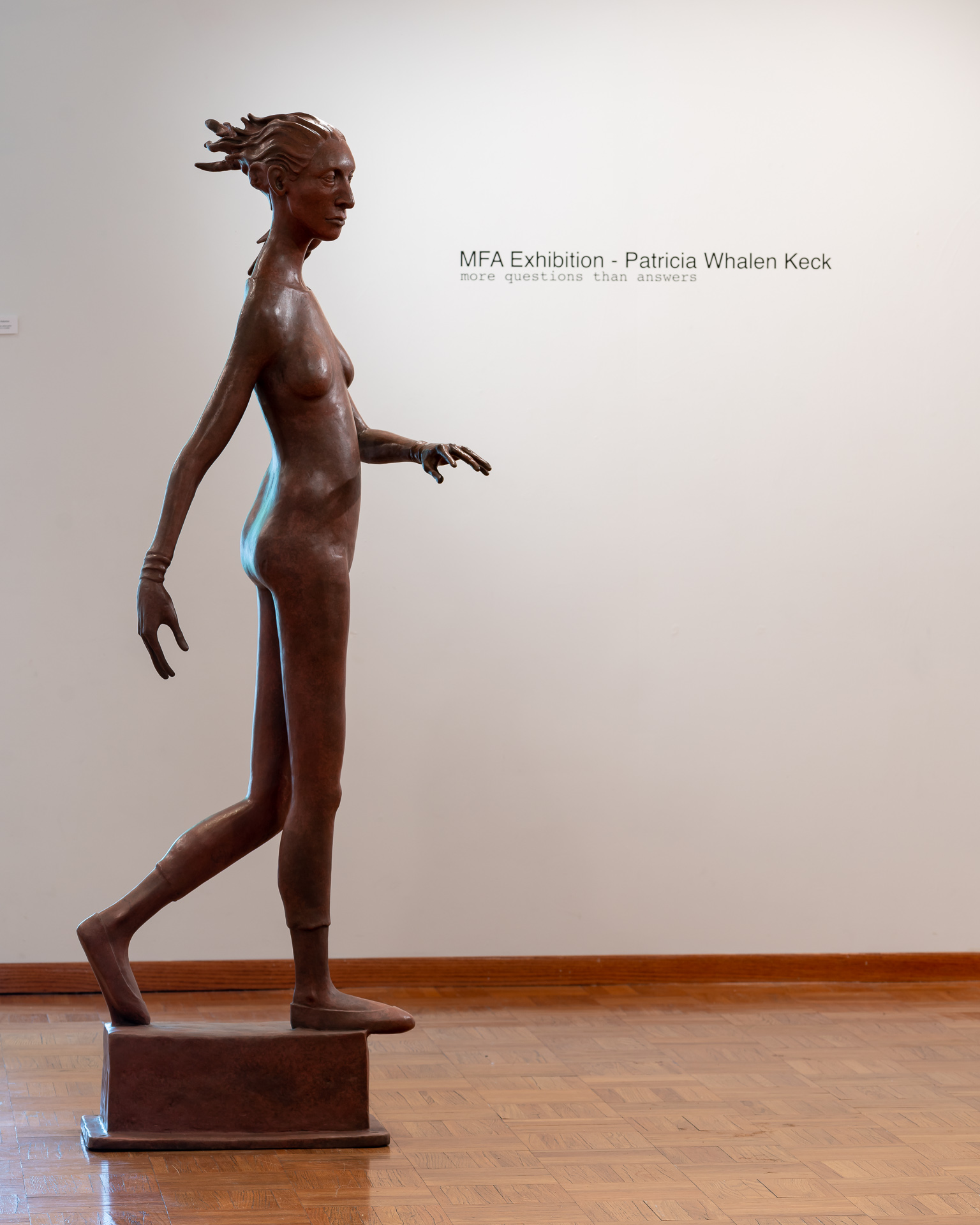 Image: Patricia Whalen Keck "The Pedestrian" Bronze. A large sculpture of a woman in a position that suggest she is walking or crossing across the space. The show title of the exhibition is seen on the wall in the background. Image courtesy of the artist