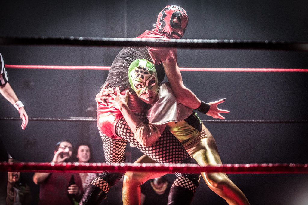 In this still from the film “Signature Move,” Zaynab (played by Fawzia Mirza) wrestles lucha libre style with Ragina Cruz (played by Molly Callinan). Zaynab faces the camera, crouching, leaning into Ragina, and gripping Ragina by the hips. Ragina’s body is angled away, with right arm outspread and hand open, as if caught by surprise. Zaynab wears a green, gold, and black lucha libre mask; a shiny, stretchy, full-length bodysuit that is gold and black; and a small silver cape. Ragina wears a red and black lucha libre mask; a black mesh and shiny red top with shiny red bottoms; and black fishnets, boots, knee pads, and accessories. In the foreground and background, the ring’s black and red ropes are visible. A few spectators cheer in the lower left-hand corner, and a referee’s arm enters the frame from the left. The frame’s edges are slightly feathered and dark.