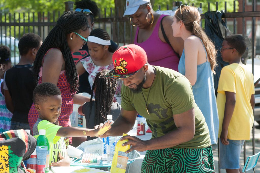 At the Bright Star block party in summer of 2018. A crowd of people stand around several tables with art materials and activities on them. The two people in the foreground are working together, holding paints, and looking down at the project they're working on. Photo by Tony Smith. 