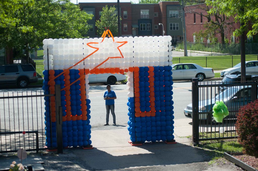 A photo from the Bright Star block party in summer 2018. There is a balloon arch made in the design of the Bright Star blue, orange, and white logo. There is a person standing just behind the threshold on the street in front of the arch. Photo by Tony Smith. 