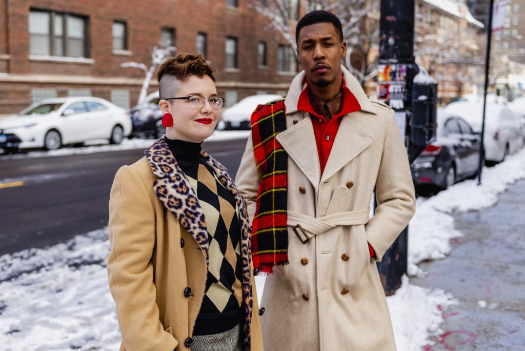 Image: Two models are seen posing on the sidewalk on 55th street outside of Gilda's. There is snow and cars in the background. The pair are looking directly into the camera. The person on the right has on a beige coat with a red-checkered scarf and red shirt. The person on the left has a brown coat on with buttons and a leopard collar. Their shirt is brown, beige, and black. Photo by Ryan Edmund Thiel.
