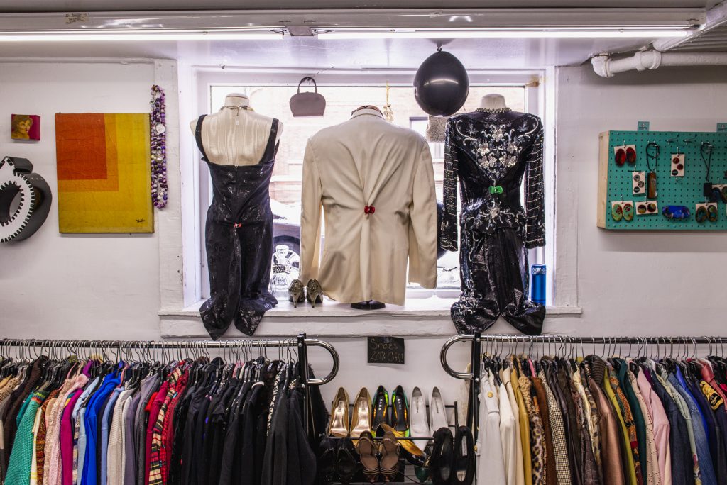 Image: A view of the inside of Gilda's store facing out towards the street. There are three mannequins in the window that are styled and displayed. From left to right, the mannequins are wearing a black jumpsuit, a cream-colored blouse, and a black and silver dress. Art and other items hang on the walls and clothes and shoes line the clothing racks on the lower half of the image. Photo by Ryan Edmund Thiel.