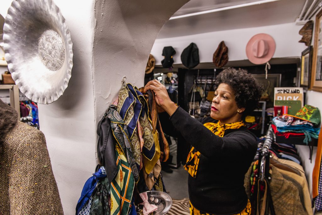 Image: A view of the interior of Gilda's, where she is seen wearing a black shirt and a yellow scarf, styling products on the wall. Several scarves hang from the walls as well as hats and other objects. Racks of clothing are in the background. Photo by Ryan Edmund Thiel.