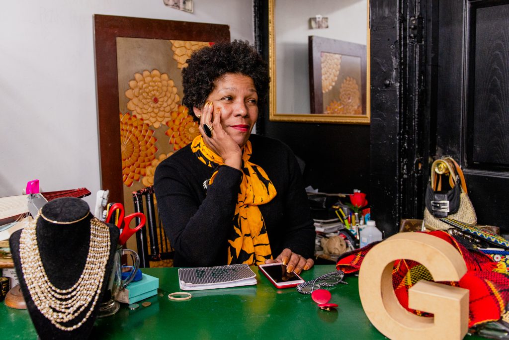 Image: Gilda Norris is seated at her desk wearing a black shirt and a yellow and black scarf. She has her chin in her hand and is looking off towards the right portion of the photo. There are items on her green desk: a wooden "G," a gold necklace, her iPhone, a notepad, pens, and scissors. In the background, there is a framed image of yellow and orange flowers and a mirror. Photo by Ryan Edmund Thiel.