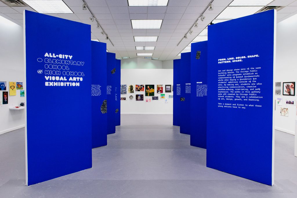 Image: Front view of the All-City High School Visual Arts Exhibition. Blue zigzag walls create a walkway for viewers with art displayed on the surrounding walls. Photo by Ryan Edmund.