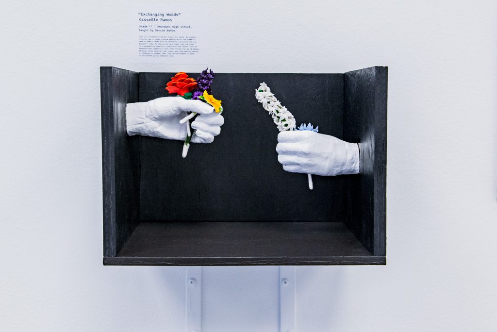 Image: Gisselle Ramos, “Exchanging Words”. A black wooden box containing white porcelain hands outreached toward one another, each holding delicate flowers. The hand on the left is positioned higher than the right. Photo by Ryan Edmund.