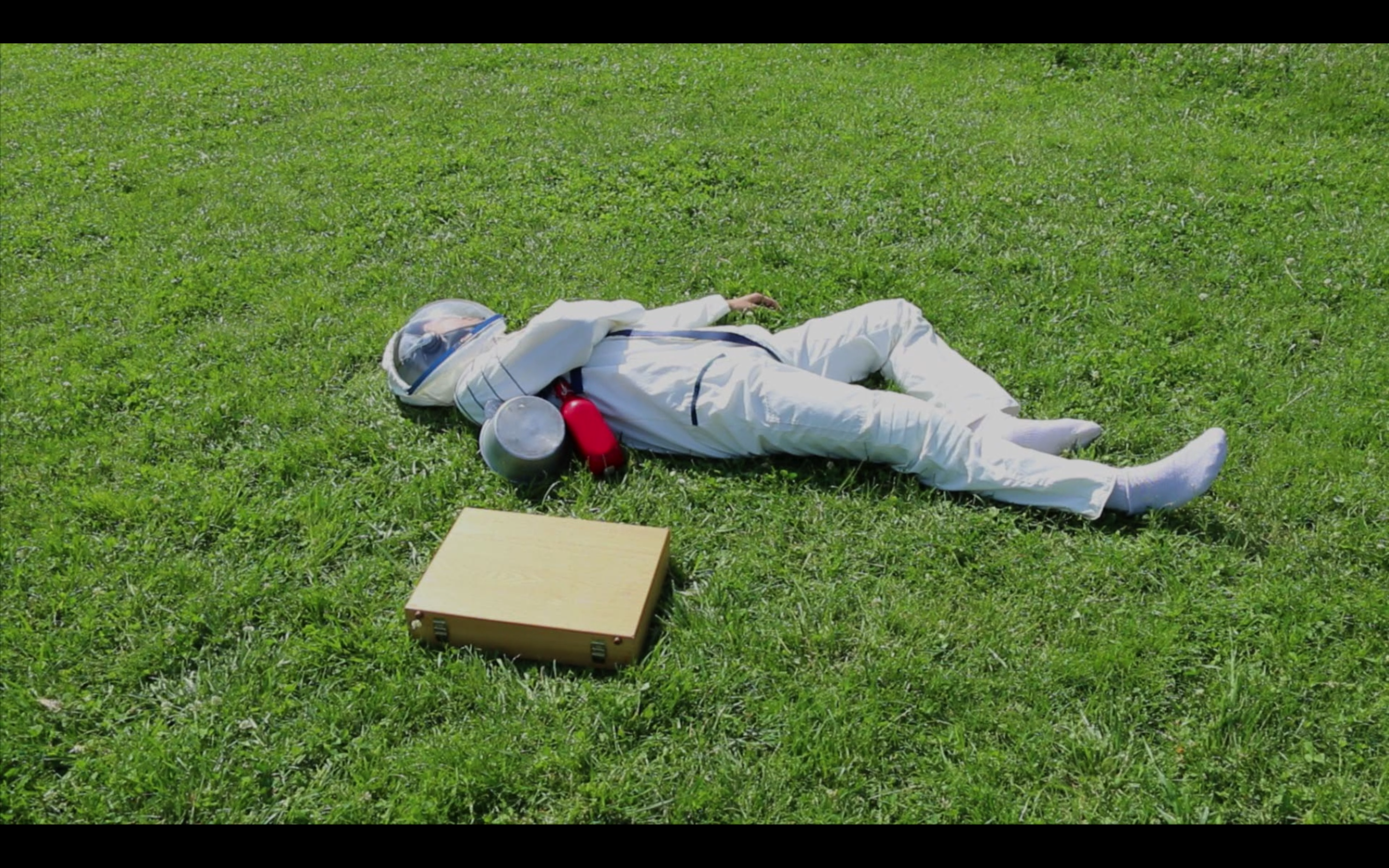 Image: A video still of a cosmonaut wearing a white space suit and helmet is laying on their back on a grassy green field with one arm across their chest. A tan briefcase is tilted on its side lays next to the figure. Image courtesy of the artist.