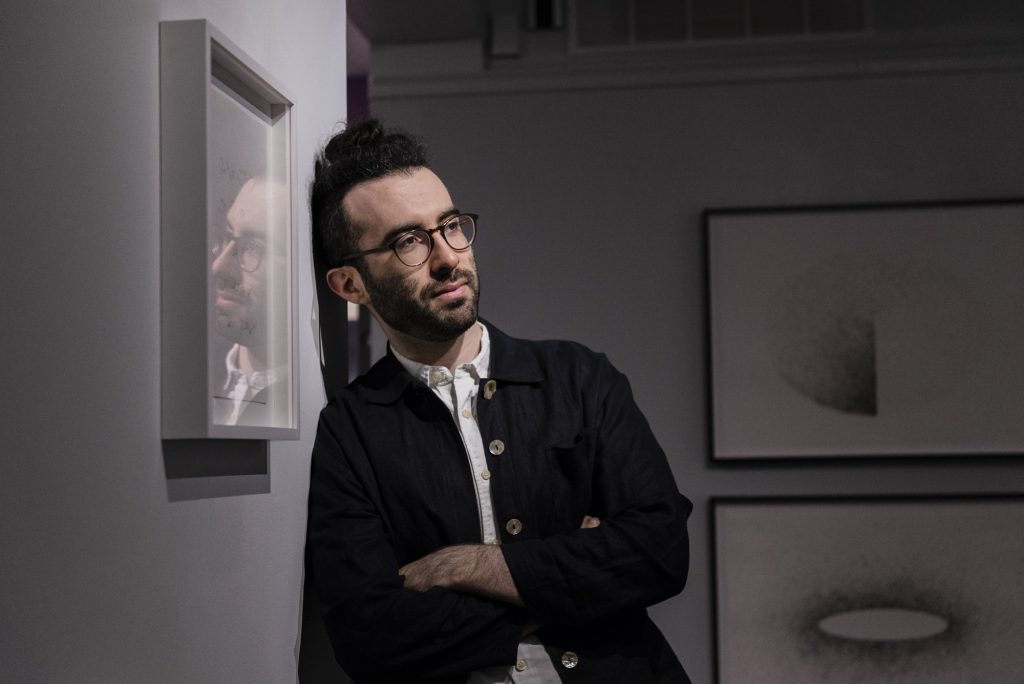 Image: The artist is leaning against a gallery wall in the LA&M. He is looking off towards the right side of the space, his arms are crossed, and he is wearing a dark button down. He has glasses and a beard. His facial reflection can be seen in a piece that is hanging on the left hand side in a frame. Two "hair drawings" can be seen, out of focus, in the back of the gallery. Photo by Ryan Edmund Thiel.
