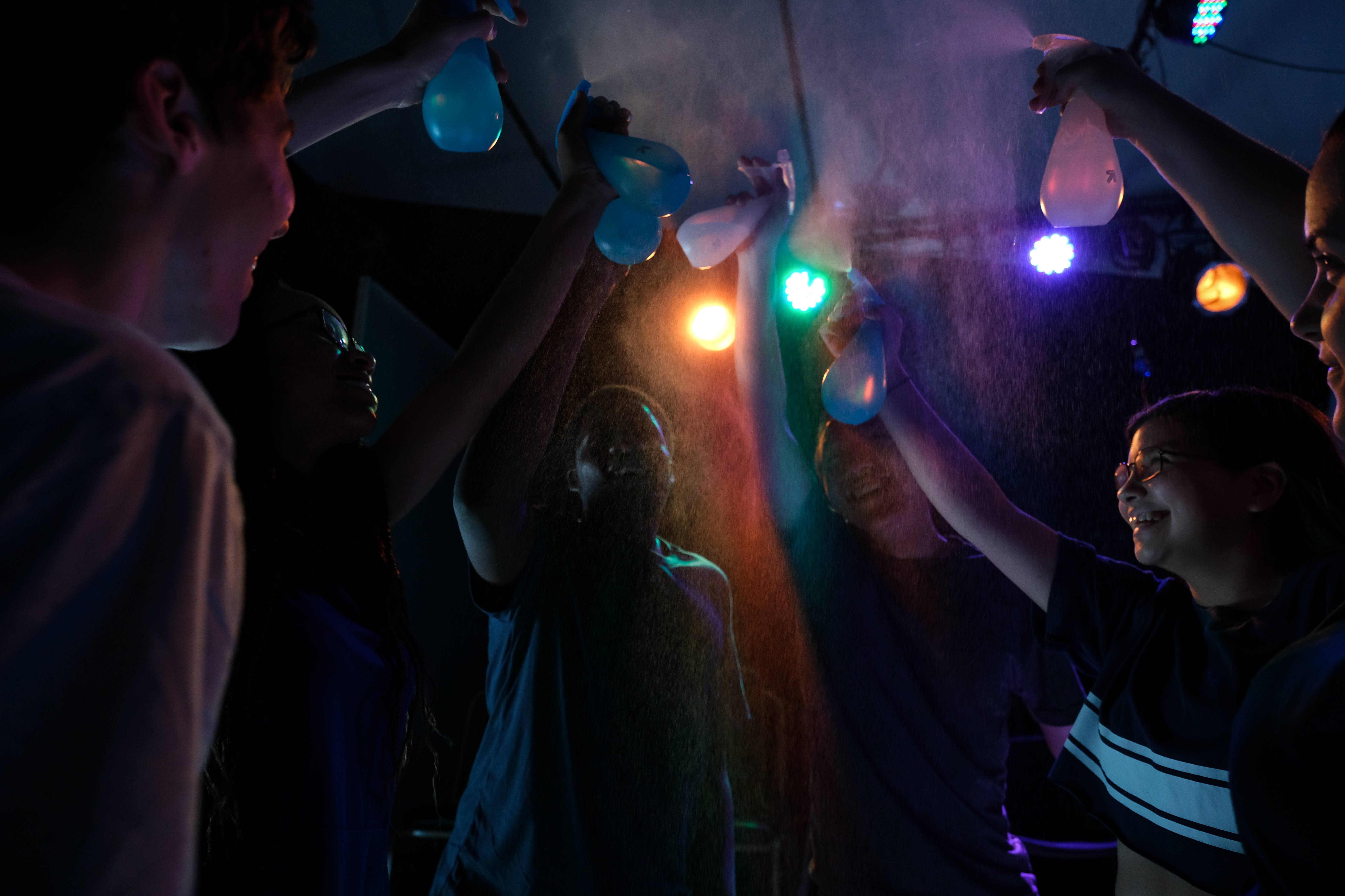 Image: Six youth performers of "Parched" circle and face in towards each other. Their arms extended and holding water bottles, they spritz the air. An orange, green, purple, and yellow lightbulb strung in the air behind them. Image courtesy of Joel Maisonet