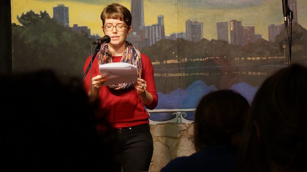 Image: Photo of Nathalie Lagerfeld performing at Miss Spoken at the Gallery Cabaret. The reader stands on-stage, speaking into the microphone and looking down at a set of papers. Lagerfeld wears a red shirt, dark pants, a multi-colored scarf, and glasses. The reader appears in medium-long-shot near the left-center of the frame. Painted on the stage wall behind the reader is a scene showing Chicago’s skyline, as if viewed from behind a stone wall. In the foreground, the backs of a few audience members’ heads are visible. Photo by Sarah Joyce. Courtesy of Miss Spoken.