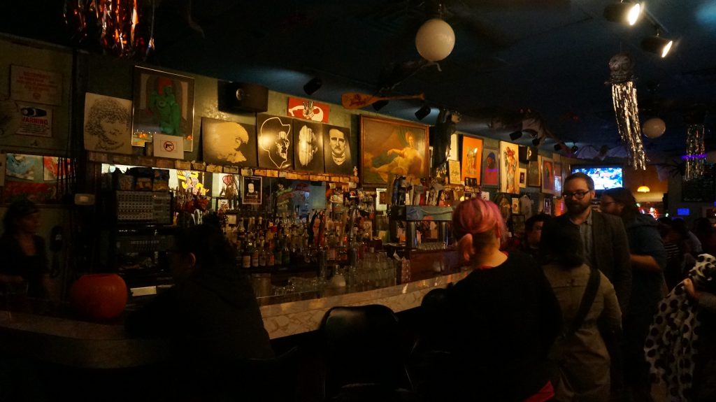 Image: Photo of the bar area at Miss Spoken at the Gallery Cabaret. Lining the wall along the back of the bar area are several drawings, paintings, and photographs. Most images show faces and/or bodies, but are otherwise diverse, reflecting a range of styles, ages, color palettes, etc. Objects like oars and metallic streamers hang from the ceiling. In the foreground, patrons mingle in the dim bar. Photo by Sarah Joyce. Courtesy of Miss Spoken.
