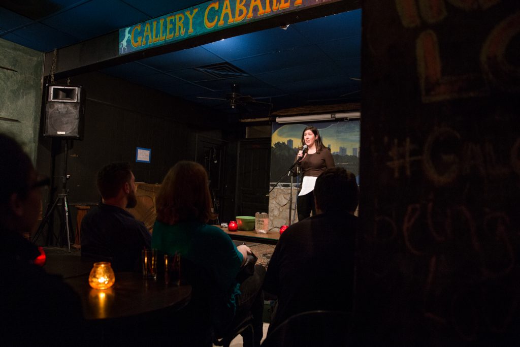 Image: Photo of Gina Watters performing at Miss Spoken at the Gallery Cabaret. The reader, wearing a dark shirt and dark pants, stands on-stage speaking into the microphone while looking out at the audience. One of Watters’ hands grips the microphone and the other holds loose papers. The reader appears small near the center of the frame. Painted on the stage wall behind the reader is a scene showing Chicago’s skyline, as if viewed from behind a stone wall. In the foreground, the words “Gallery Cabaret” are partially visible at the top of the frame. The backs and sides of some audience members’ heads are visible at the bottom of the image. Photo by Sarah Joyce. Courtesy of Miss Spoken.