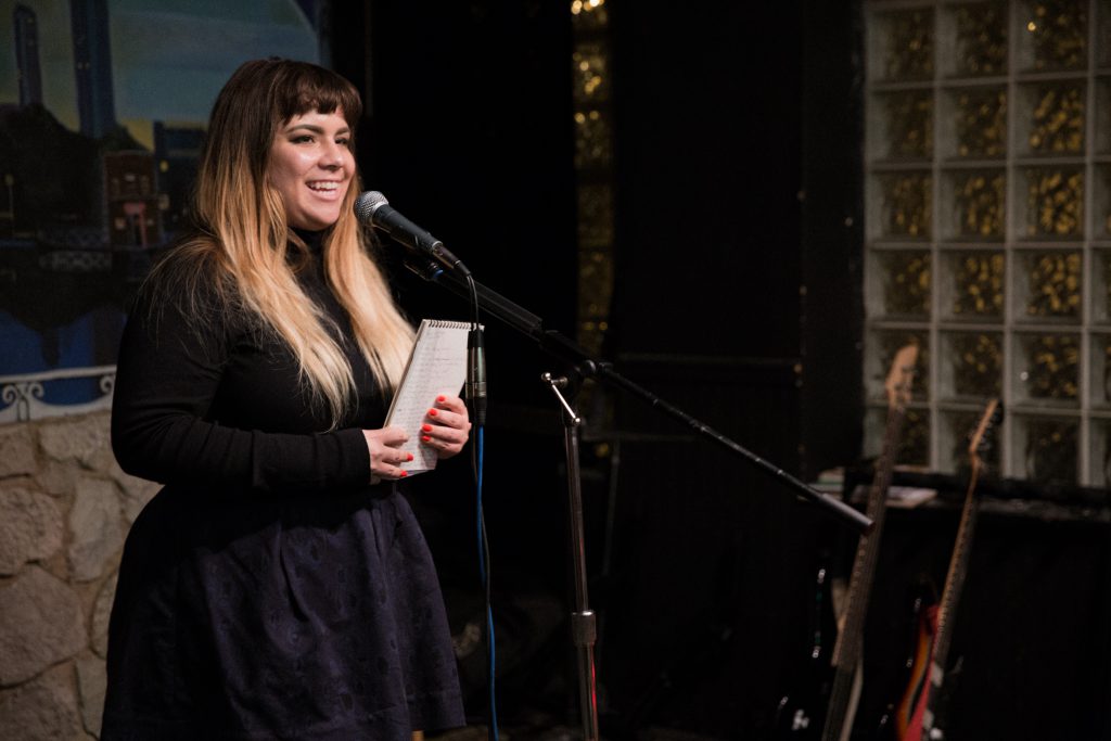 Image: Photo of Gina Wynbrandt performing on-stage at Miss Spoken at the Gallery Cabaret. Wynbrandt smiles out toward the audience (not visible) and holds a spiral notebook in both hands. Wynbrandt wears a black shirt and navy skirt. Painted on the stage wall behind the reader is a scene showing Chicago’s skyline, as if viewed from behind a stone wall. Also visible in the background are two electric guitars, set to lean toward a black wall with glass-brick windows. Photo by Sarah Joyce. Courtesy of Miss Spoken.