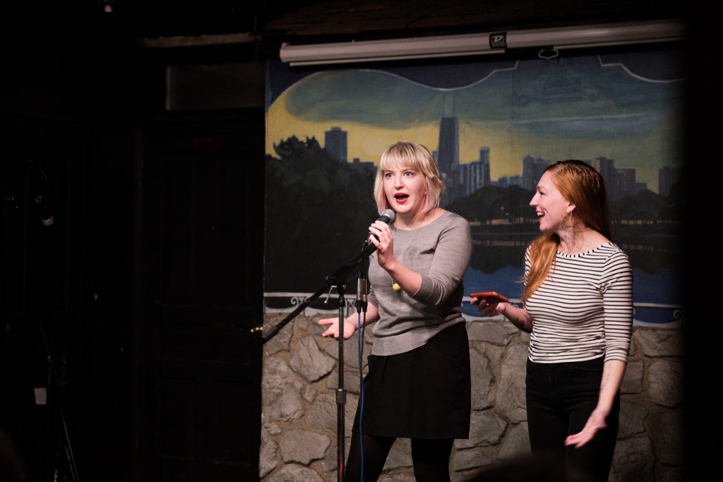 Image: Photo of Rosamund Lannin (left) and guest co-host Alicia Swiz standing on-stage at Miss Spoken at the Gallery Cabaret. Both appear in medium-long-shot on the right side of the frame. Lannin looks out toward the audience (not visible) and speaks into the microphone, while Swiz smiles and looks at Lannin. Lannin wears a grey shirt, black skirt, and black leggings. Swiz wears a black and white striped shirt and black pants. Painted on the stage wall behind the readers is a scene showing Chicago’s skyline, as if viewed from behind a stone wall. Photo by Sarah Joyce. Courtesy of Miss Spoken.