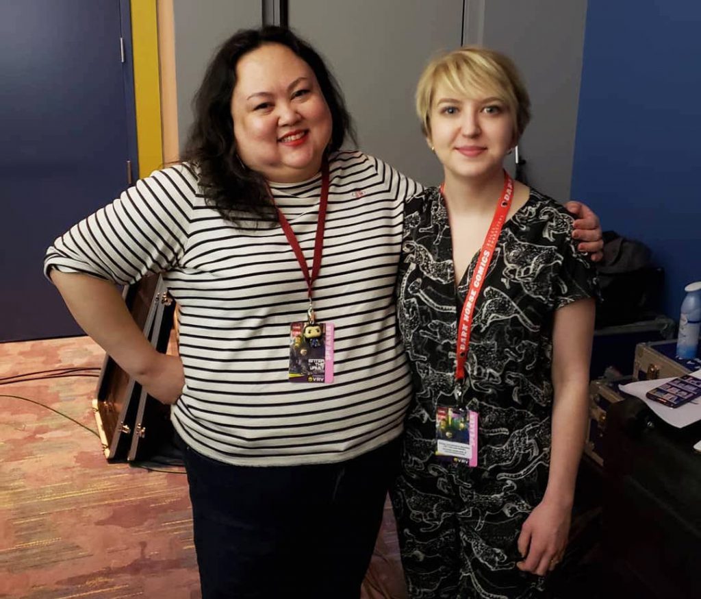 Image: Jasmine Davila (left) and Rosamund Lannin (right) stand together backstage at C2E2 for Cards Against Humanity Theater, smiling for the camera. Davila wears a white and black striped shirt and black pants, and Lannin wears a patterned black and white jumper. Both wear red lanyards around their necks and badges that read “Speaker.” Davila has one arm around Lannin’s shoulders. Around them backstage are A/V equipment cases and miscellaneous objects. Courtesy of Miss Spoken.