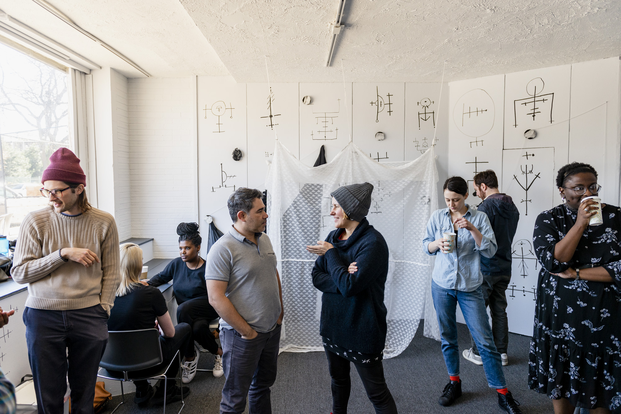 Image: A group of people standing inside Roman Susan. Zuri Washington (far right) transformed the space by painting black sigils on the white walls and adding fiber elements. Event co-curator Adia Sykes (back left) is sitting at a table with event attendee and facilitating a tarot card reading. Photo by Ryan Edmund.