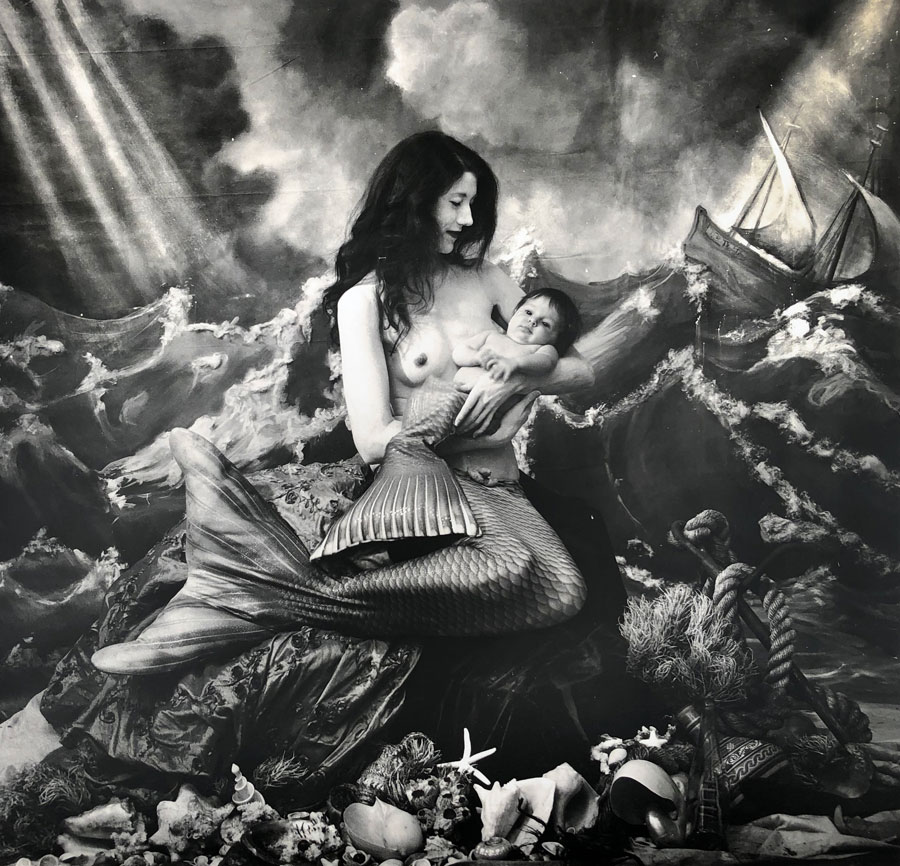  Image: A black and white photograph of a woman wearing a mermaid tail, seated on a seashell-ladened formation. She is bare-chested holding a baby, also adorned with a mermaid tail. Behind them is a painted backdrop depicting a sea full of waves and a ship off to the right. A Mermaid’s Tale, NM, 2018 © Joel-Peter Witkin / image courtesy Catherine Edelman Gallery, Chicago. 