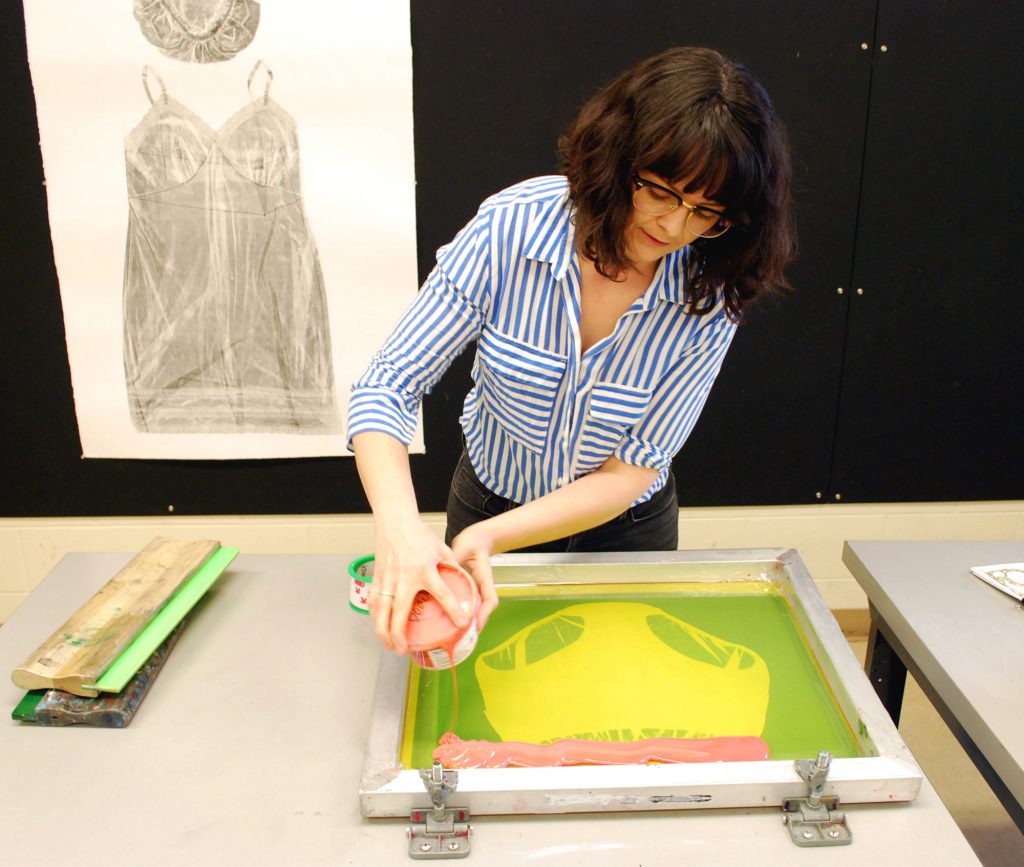 Image: Guen Montgomery pours neon pink paint onto a screenprinting screen with an image of women’s underpants on it. Pinned behind her on a black corkboard is a gray collagraph print of a woman’s shower cap and nightgown on white paper. Photo by Jessica Hammie.