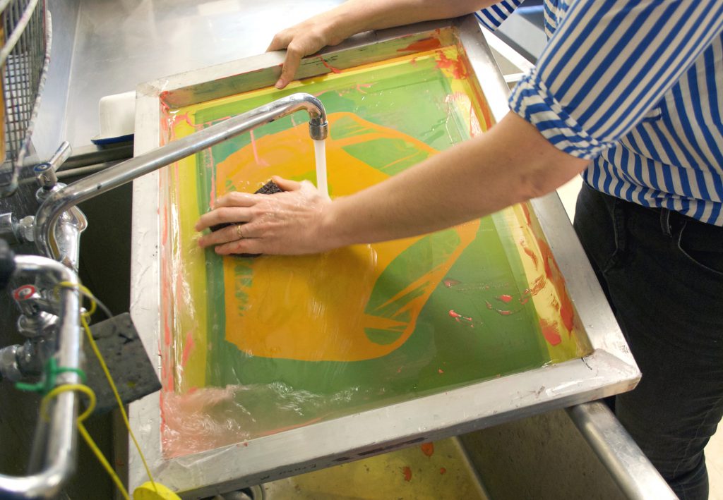 Image: Guen Montgomery washes a screenprinting screen in a small sink. Her left hand uses a sponge to wash of neon pink paint as water pours out of a faucet and onto the screen. Photo by Jessica Hammie.
