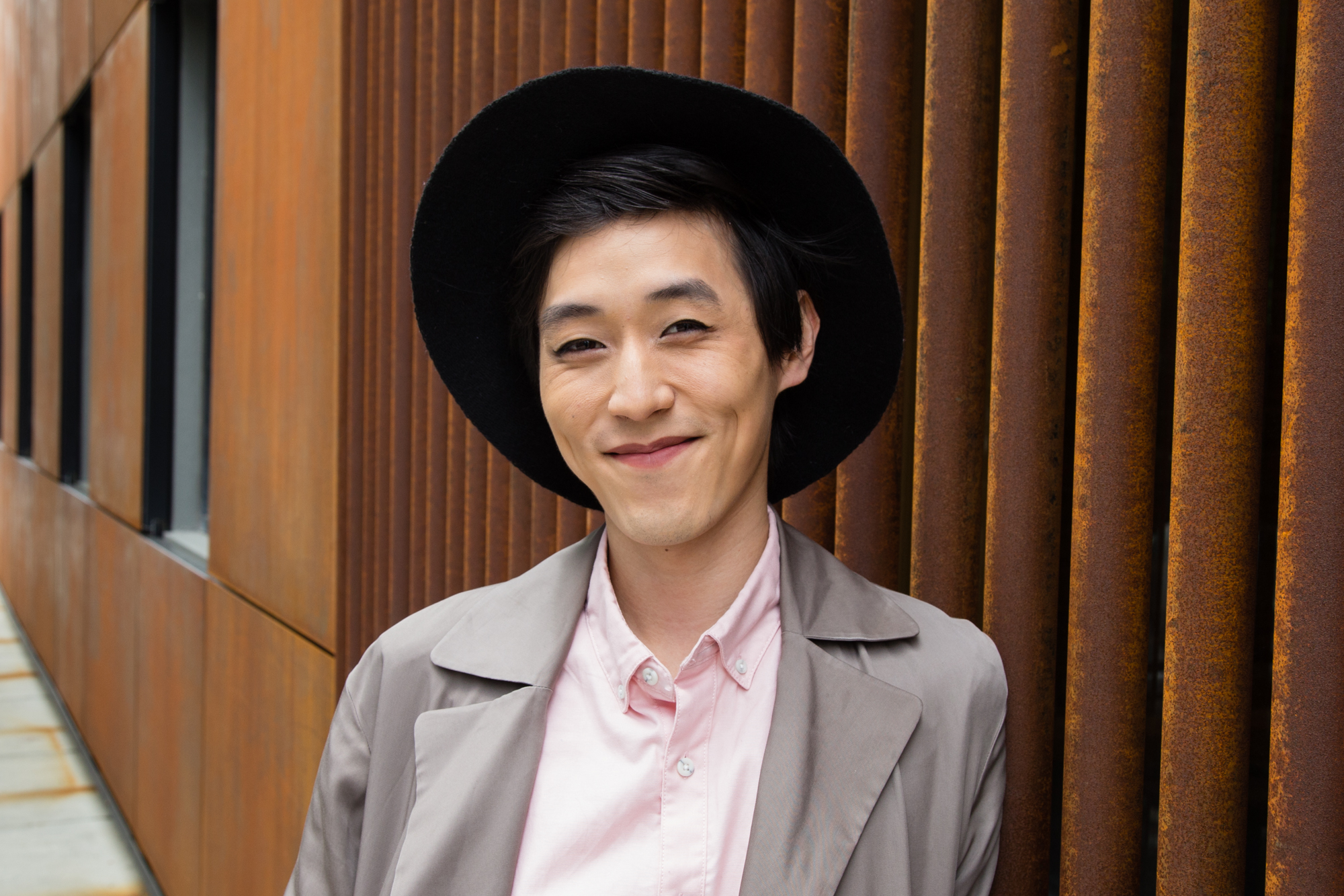 Image: Portrait of Gavin Pak leaning on a rust colored building. Gavin wears a black hat that is pushed back so their entire face is visible. A grey trench coat covers their pink button-up shirt. Photo by Joshua Johnson