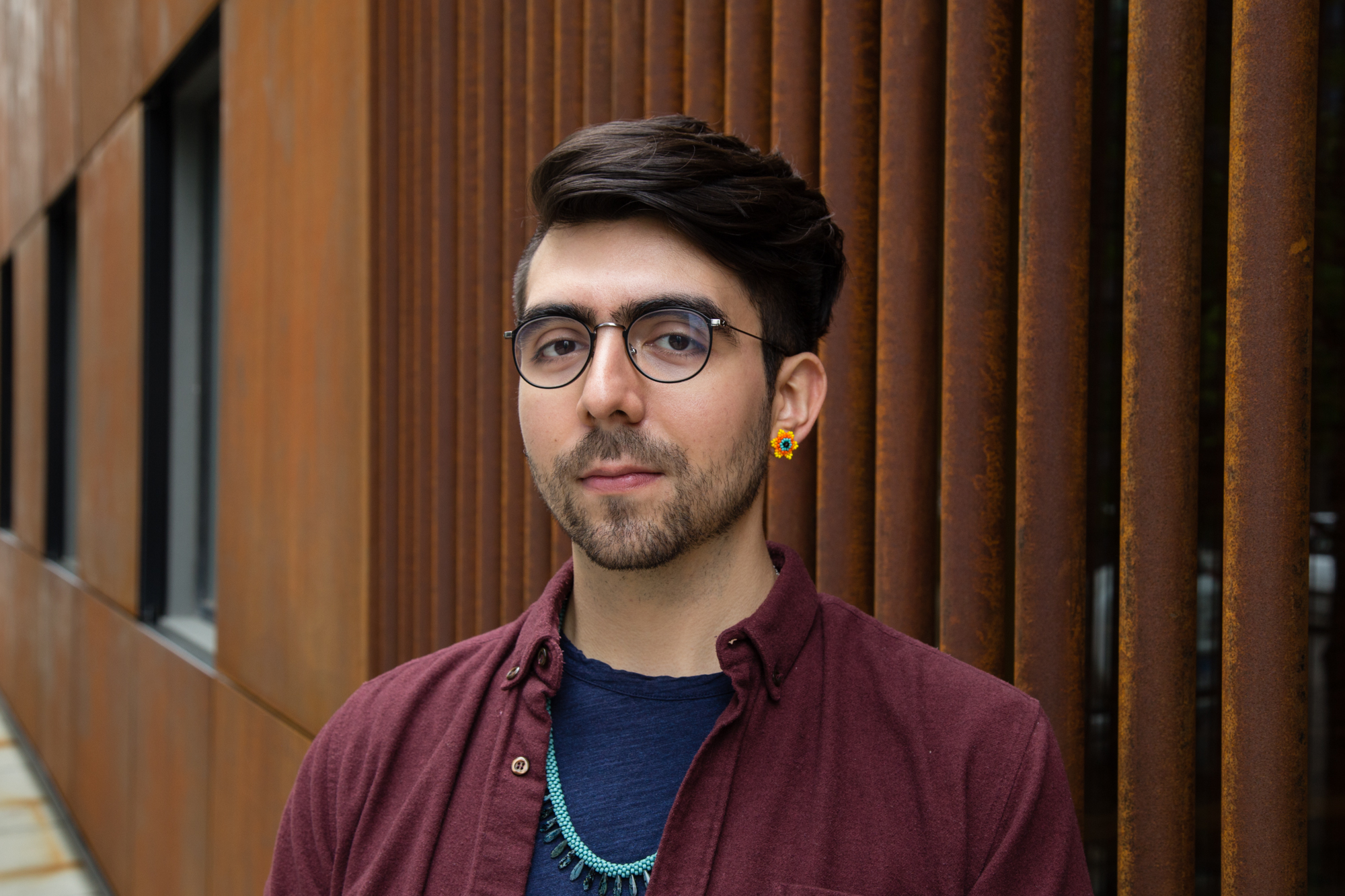 Image: Portrait of Lucas Garcia standing in front of a rust colored building. Lucas’ body is angled to the left slightly and they wear a maroon button up with a navy t-shirt underneath. On their left ear is a golden flower earring.  Photo by Joshua Johnson.