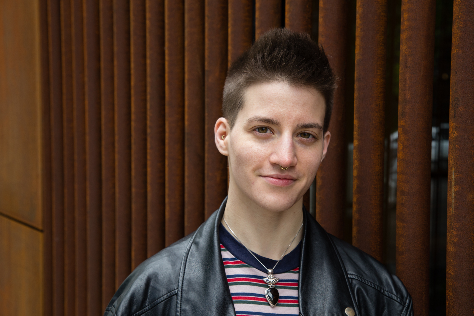 Image: Portrait of Theo Germaine in front of a rust colored building. Theo is wearing a striped pink, red, and blue shirt underneath a black leather jacket. They have a small nose-ring and wear a silver necklace with a large black stone. Photo by Joshua Johnson