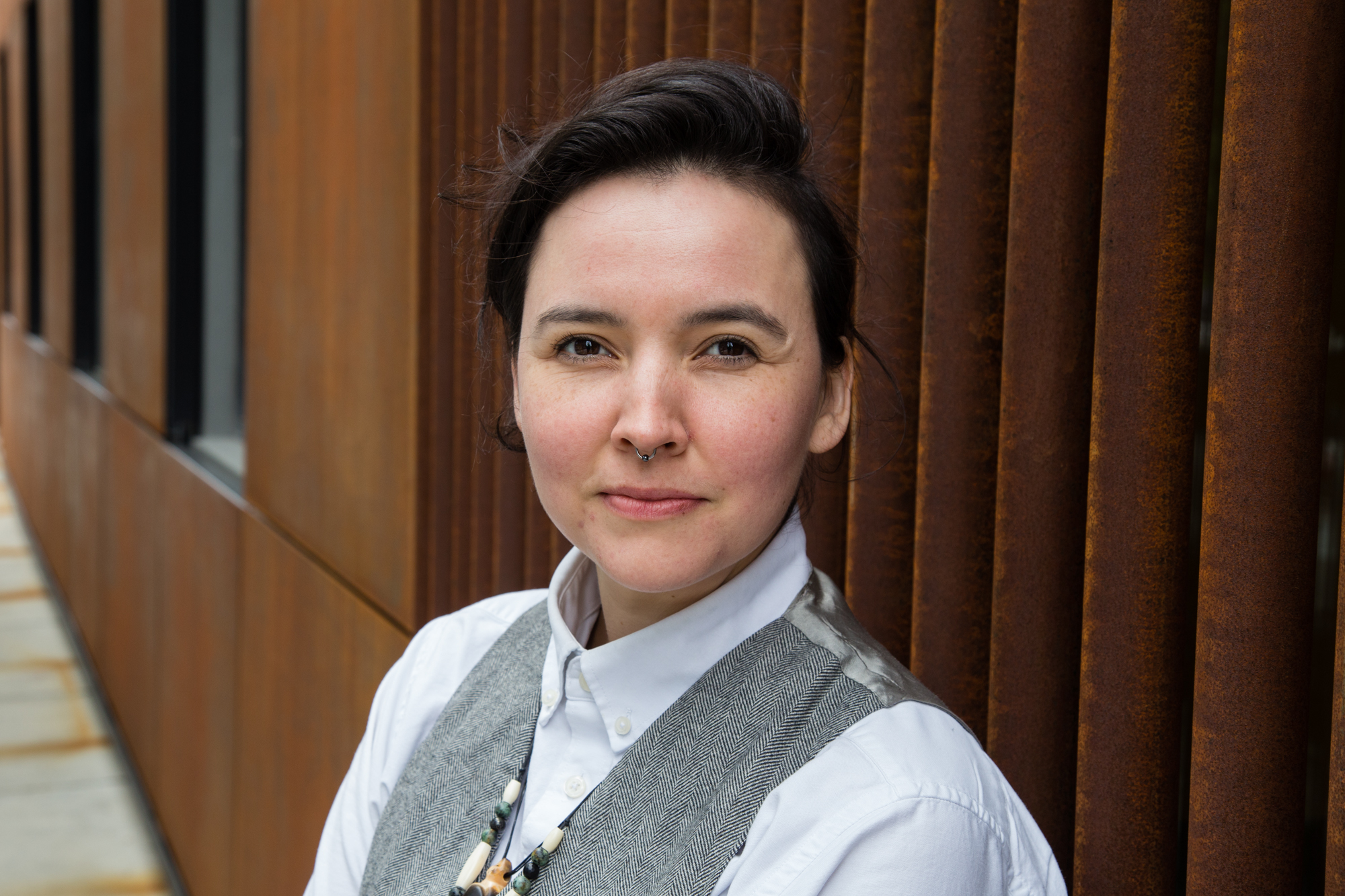 Image: Portrait of June Thiel in front of a rust colored building. June’s body is angled away from the photographer with their head facing forward. They have a small silver nose ring and wear a grey suit vest over a white buttoned collared shirt. Photo by Joshua Johnson