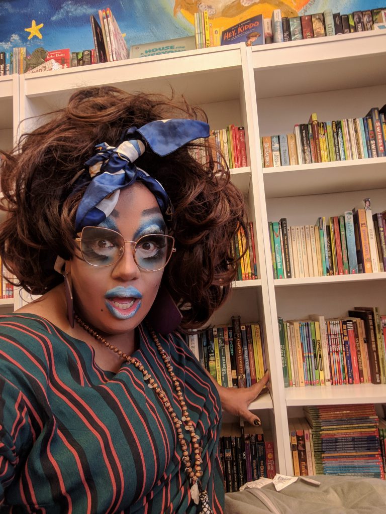 Image: Artemisia VanHo in front of a colorful bookshelf at Lit on Fire, a local bookstore in Peoria IL. Photo by the author.