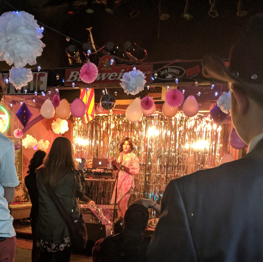 Image: Peoria Based Drag Queen Edie La'Conte Performing at Girl Party, an event put on in Peoria IL. She is shown on a stage performing under bright warm lighting, with gold tinsel and decorations put up in great amounts. Photo by the author.
